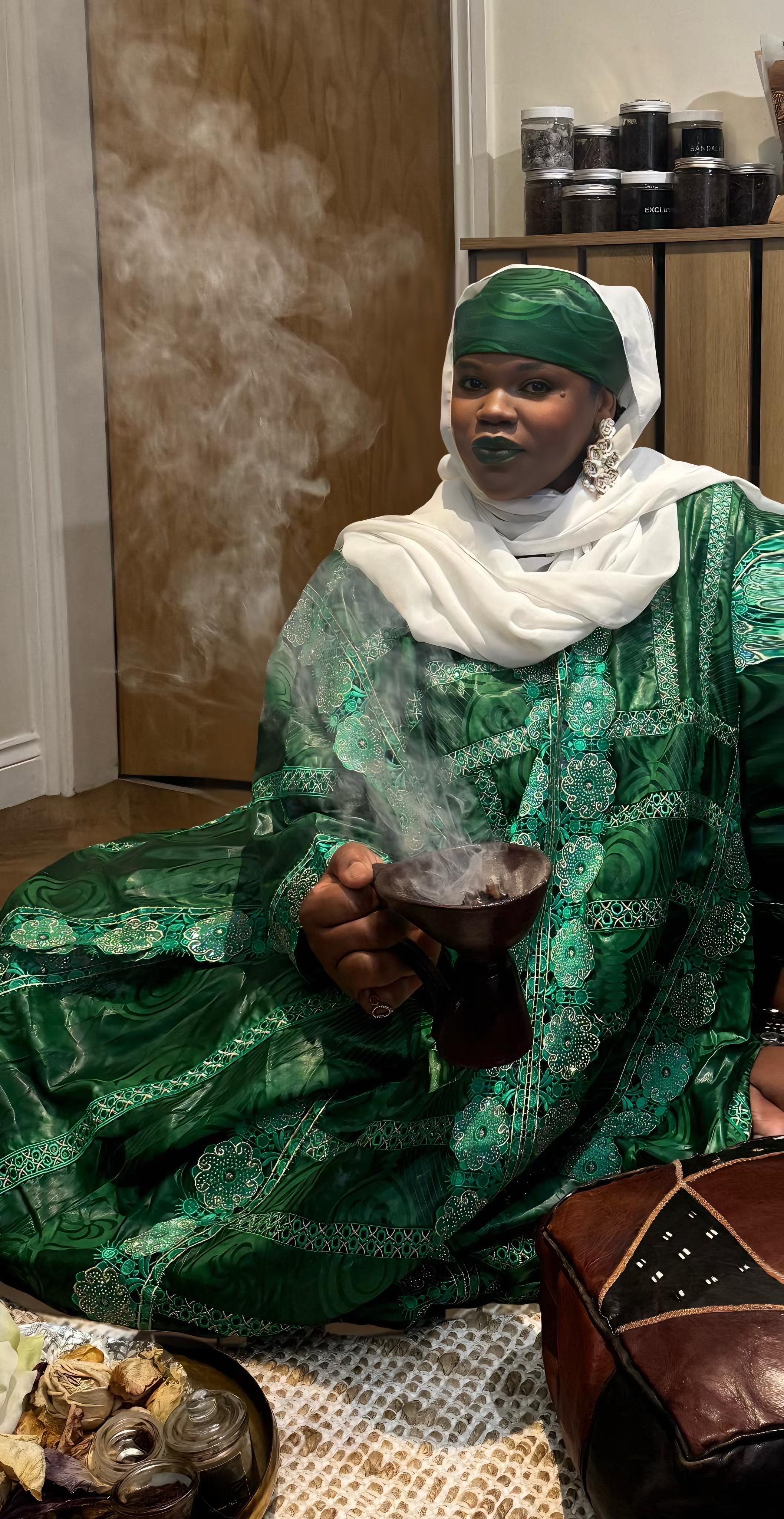 Woman in traditional green attire holding incense burner with smoke, representing Sahel wellness rituals of natural oils, herbs and self-care.
